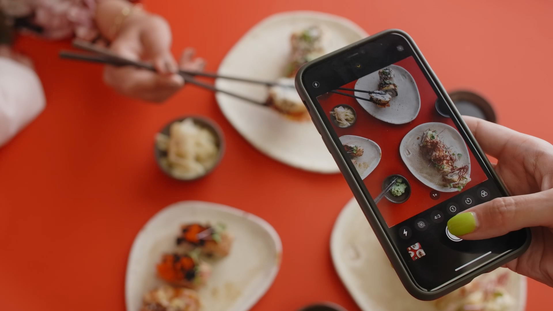 A person holds a cell phone over a table full of food to take a picture. The image on the phone is in focus but the food itself is not.